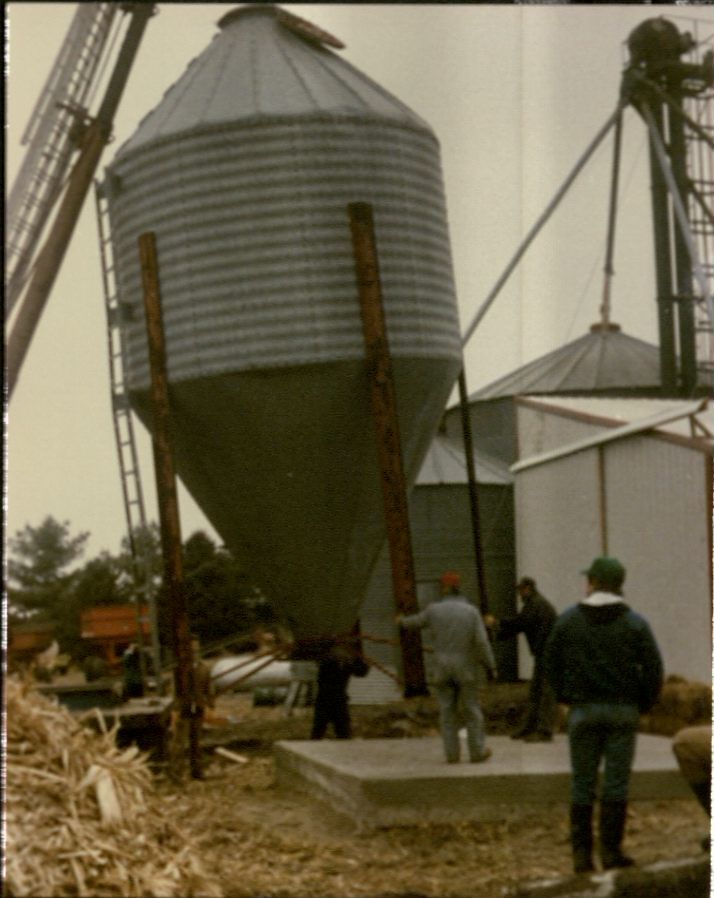 Color snapshot of a crane setting a small grain bin up on a cement platform. People are standing around and helping guide it into place.