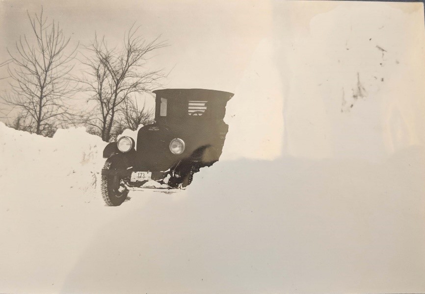 Black-and-white photograph of car driven on snowy road, snow banks in foreground, trees in background.
