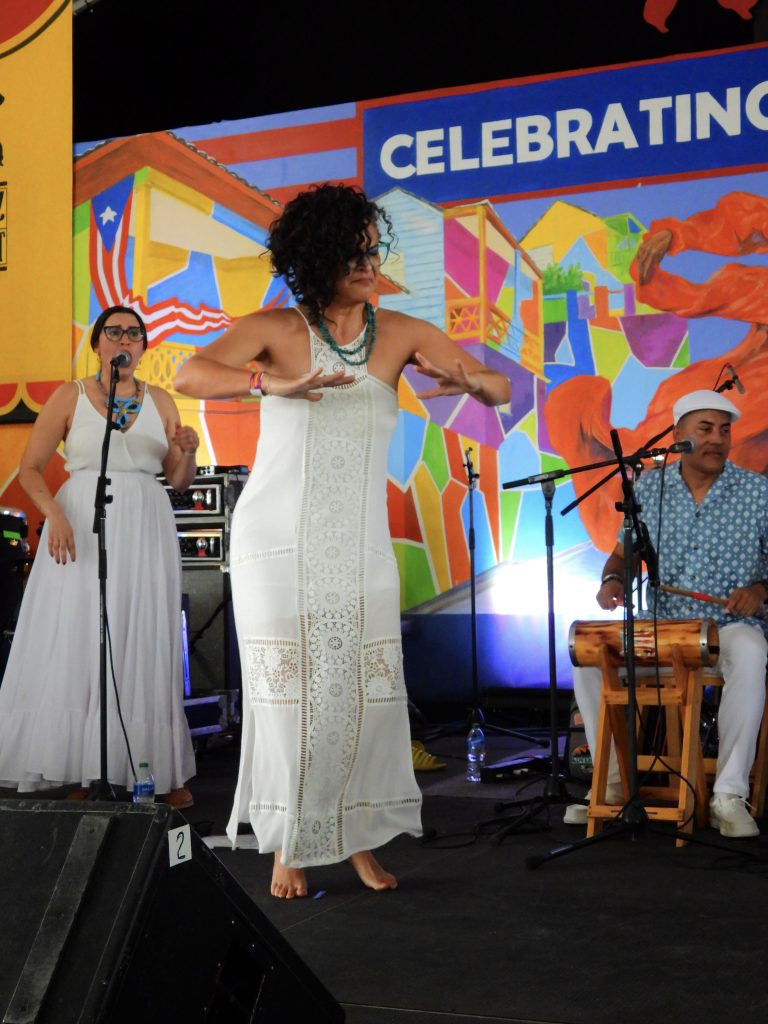 A female performer dancing Bomba in front of two musicians and a colorful background