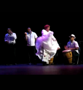 Mujer bailando Bomba en el escenario al frente de un conjunto de músicos.