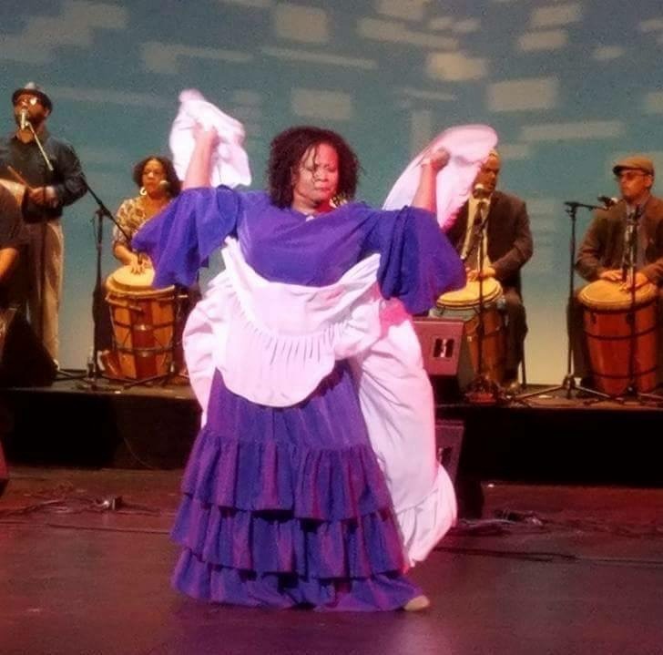 Una mujer vestida en un traje de Bomba púrpura en un escenario con una banda tocando detrás