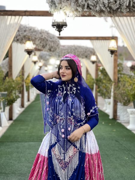 Woman posing for the camera outside. She is wearing a colorful outfit including a full skirt, a fitted top with embroidery, and a see-through, light-weight shawl.
