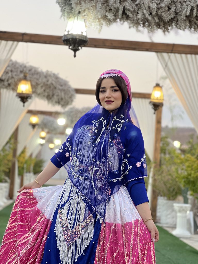 Woman posing for the camera outside. She is wearing a colorful outfit including a full skirt, a fitted top with embroidery, and a see-through, light-weight shawl.