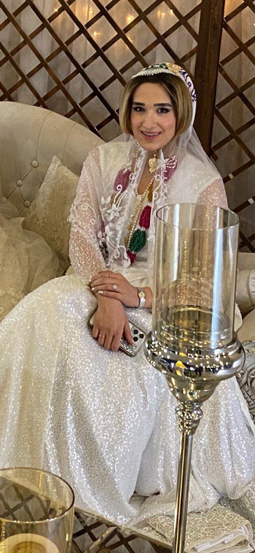 Woman sitting on a couch. She is wearing a floor-length skirt and light-weight shawl around her shoulders.