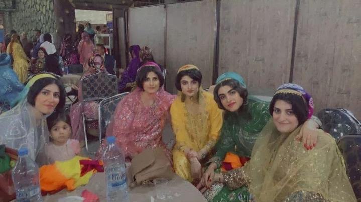 Five adult women and one child sitting around a circular table facing the camera for a picture. The women are all wearing different colors and variations of the same outfit which includes a fitted top with a light-weight shawl over top.