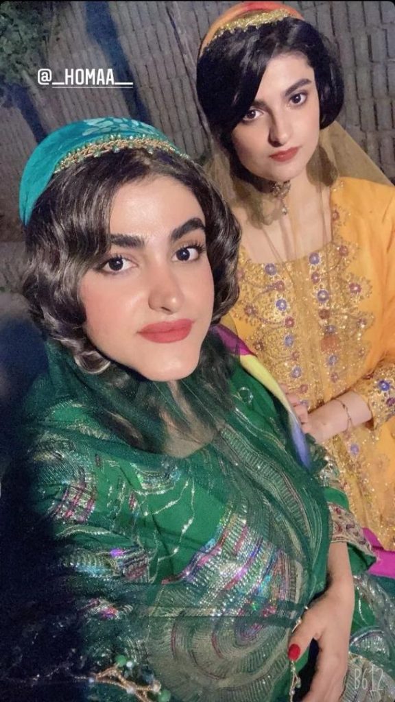 Two women taking a selfie. They are each wearing a variation of the same ensemble with a fitted top that features sequins and embroidery all over it. They also wear a see-through, lightweight shawl. On their heads is a hat that sits at the back of their head.