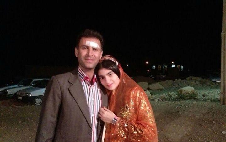 Man and woman posing for a picture outside during the night time. The woman and man are close. The man is wearing a suit jacket and button up shirt. The woman is wearing a fitted top with sequins adorned all over it. Over the fitted top she is wearing a light-weight shawl.