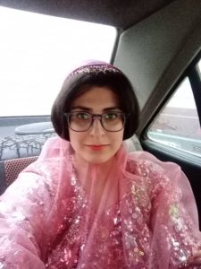 Woman sitting in a car taking a selfie. She is wearing a light-weight, fitted top with sequins.