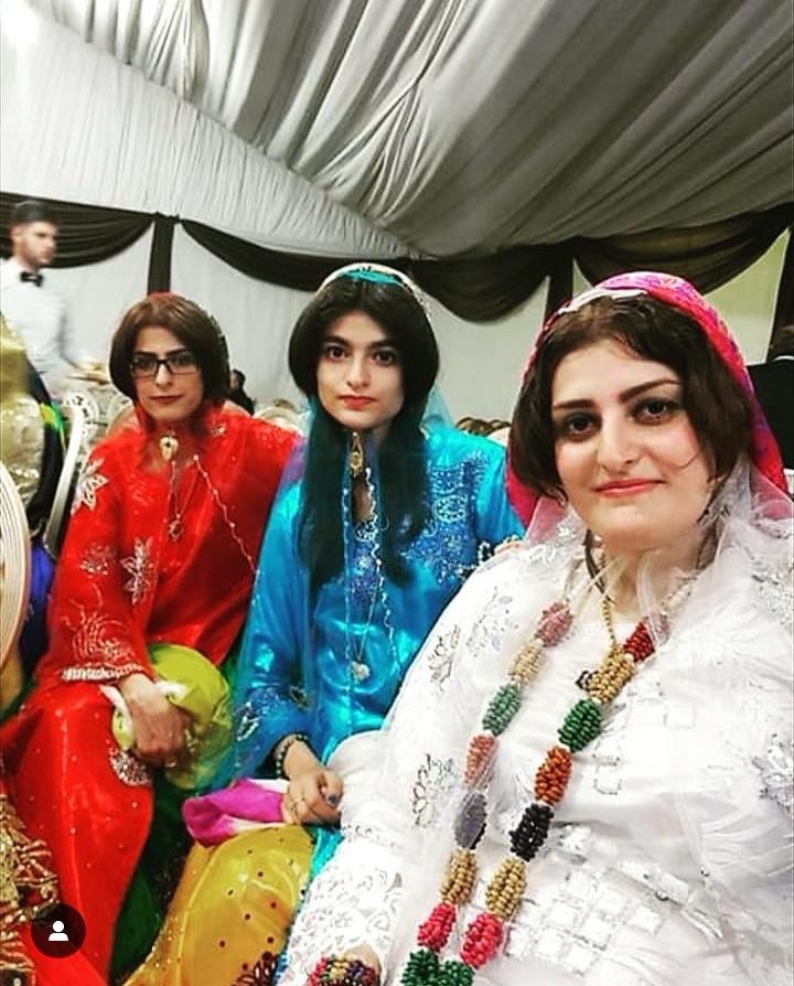 Three women attending a wedding. They are sitting and facing the camera, each with a slight smile. They are inside a tent made of fabric where the wedding events took place. They are each wearing a full-length skirt, a light-weight, fitted top, and their ensembles have sequins attached to them in various geometric and floral patterns.