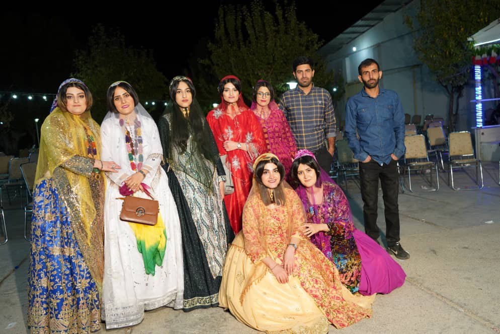 Group of 9 people posing for the camera. There are 7 people standing in a line and then 2 people kneeling in the front row. The 7 women in the photo are all wearing floor-length skirts, a light-weight shawl of varying colors. The two men pictured are wearing button-down shirts with pants.