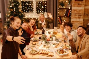 A group of people gathering around a table full of food in a festively decorated room, waving and smiling at a phone being held up at the end of the table.