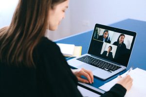 A person with long hair and wearing a suit is on looking at a virtual call on their laptop, where two other people dressed in similar working outfits look back at them.