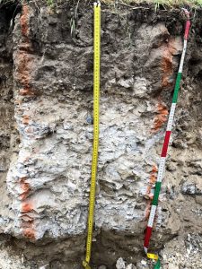 This soil profile has grey-brown soil colors and a little vegetation at the surface, mid-soil is white and appears to be like rock, at 80 cm, the soil turns back to brown and the rock-like appearance of the cemented calcium carbonate disappears