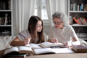 Photo of an older woman and girl sitting at a table. The older woman is helping to teach the girl who is studying.