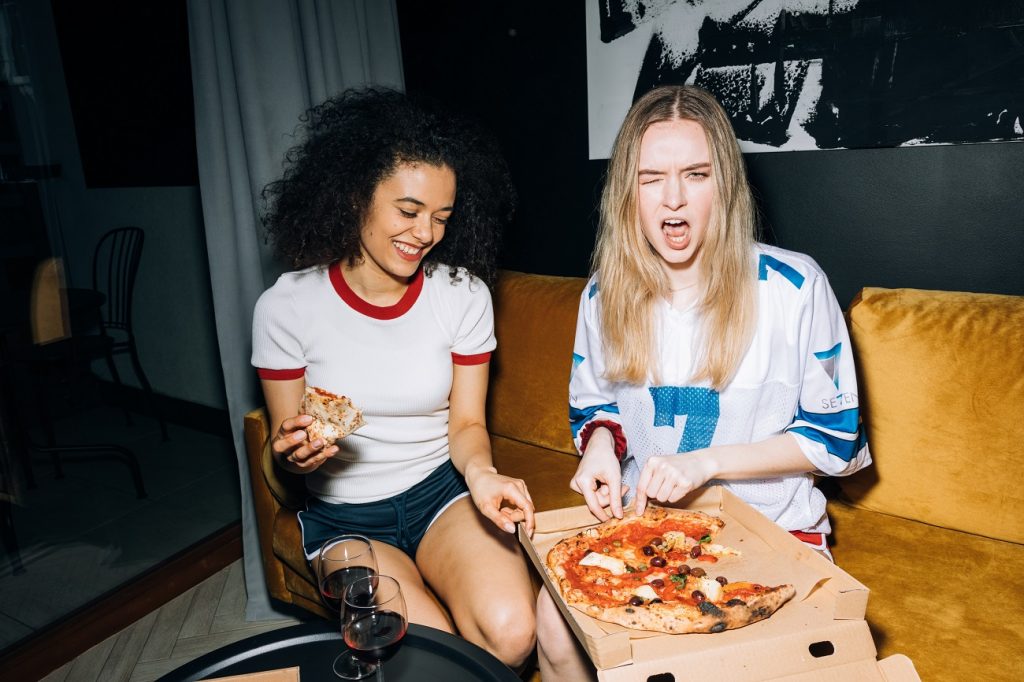 Two teenagers lounging in sportswear and eating pizza in a dark room.