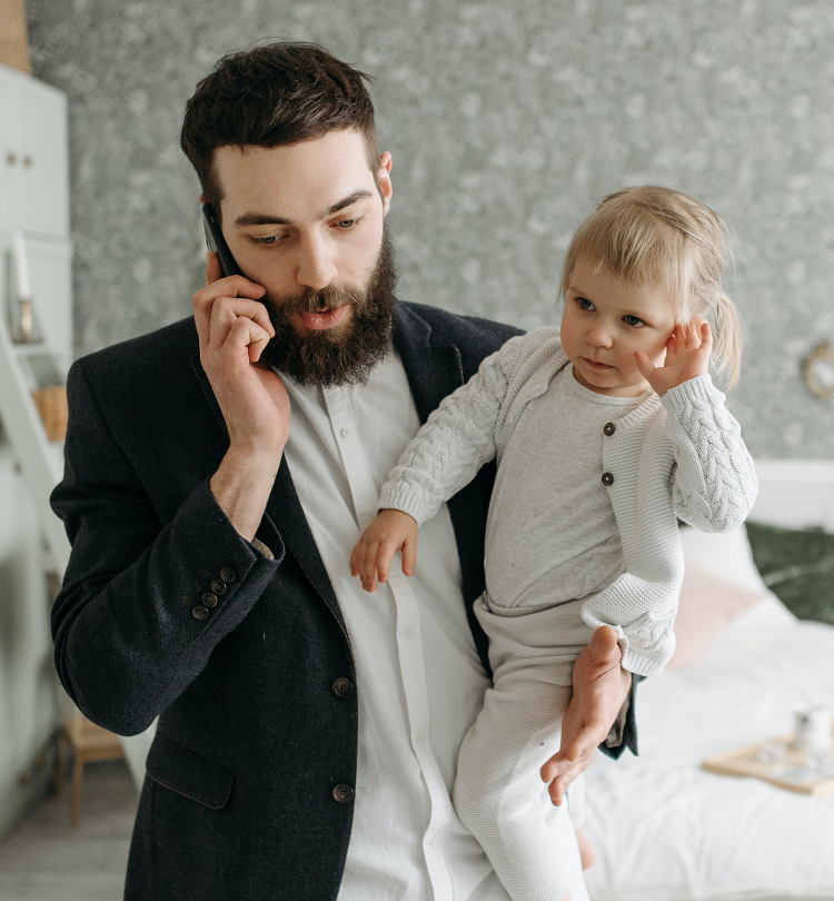 A man in a suit holds a toddler in one arm while speaking on a cell phone.
