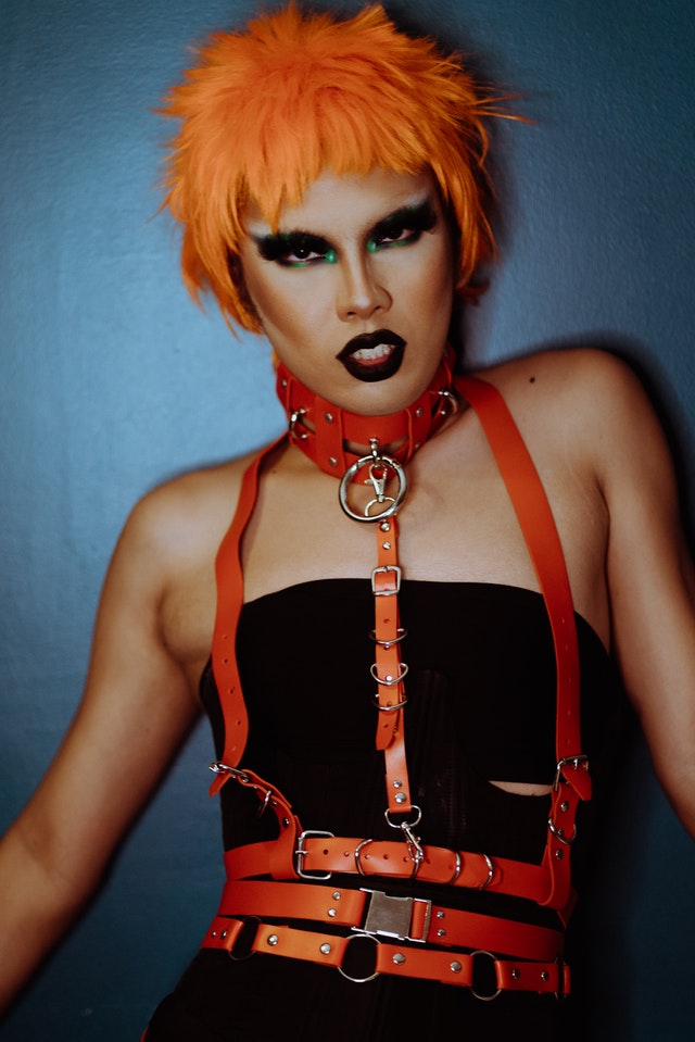 A model posing in BDSM gear, including a bright orange body harness, dark makeup, and a choker collar over tight black underclothes.