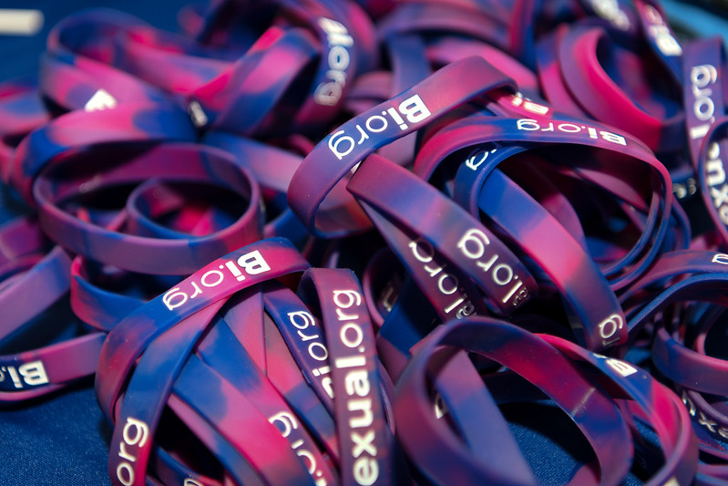 A pile of plastic arm bands with pink, blue, and purple colors mixed and bi.org printed on the front.