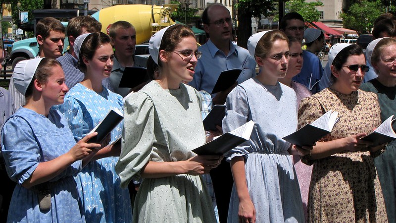 A group of Mennonite women with starched caps and conservative, flower patterned dresses, singing out of books outdoors. Mennonite men stand behind them, also singing.