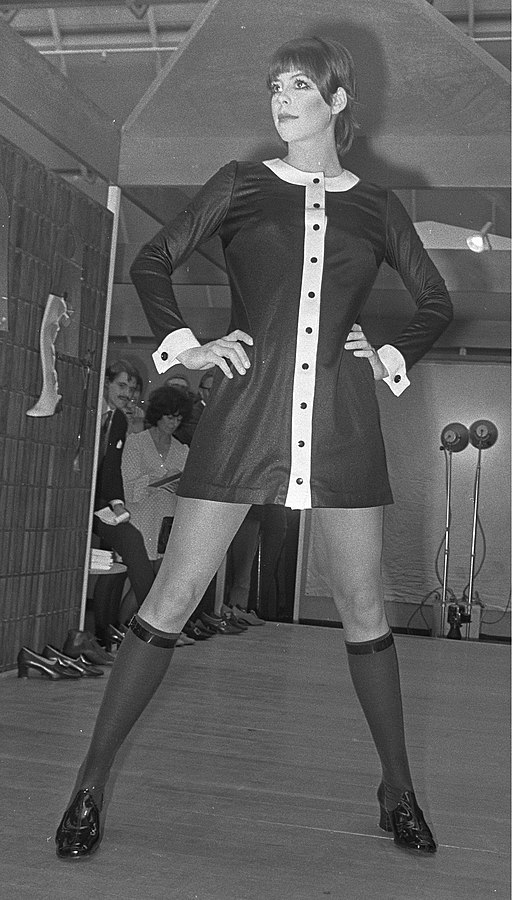 A black and white photo of a model in a short dress with knee high stockings.