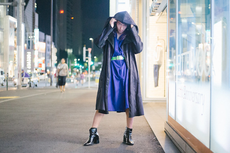 A young Asian person wearing a purple dress, black boots, and a hooded coat. They are bald and appear masculine.