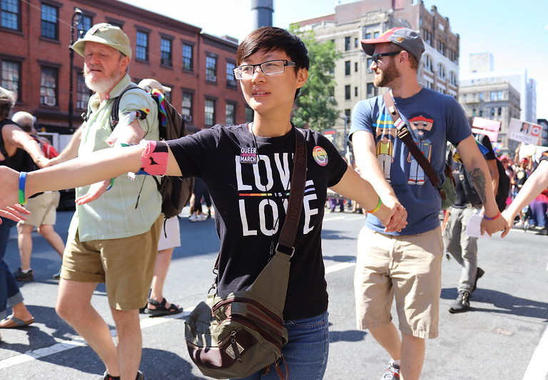 A young asian person links arms with other members of a March in New York, wearing a "Love is love" tshirt, various pins, and rainbow arm bands and bracelets.