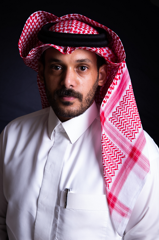 A man wearing a white button up shirt, red patterned ghutra, and a black igal holding the ghutra in place.