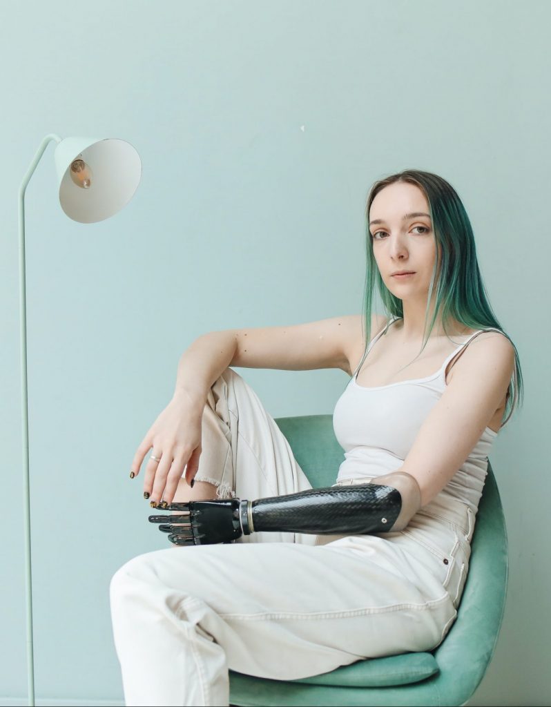 A woman wearing all white sits with a black prosthetic arm lying casually across her lap.