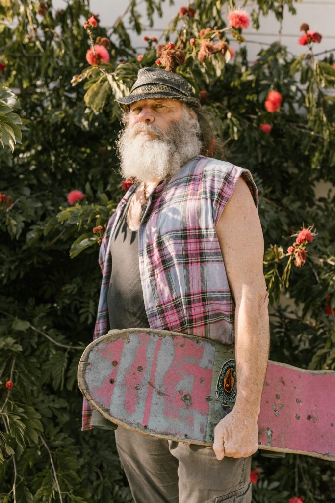 An older man with a beard, holding a tattered skateboard and wearing muted colors. His clothing is described in the caption.