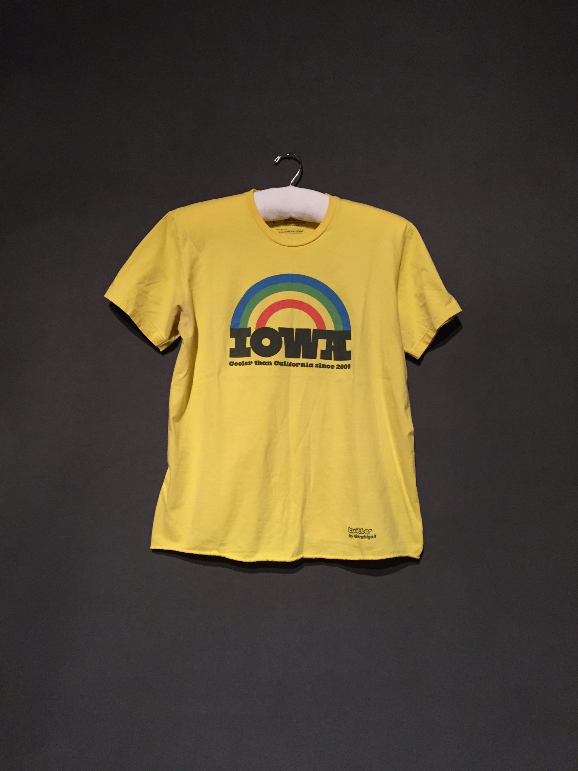 Yellow short sleeve t-shirt with a rainbow and black text reading: "Iowa: Cooler Than California Since 2009"