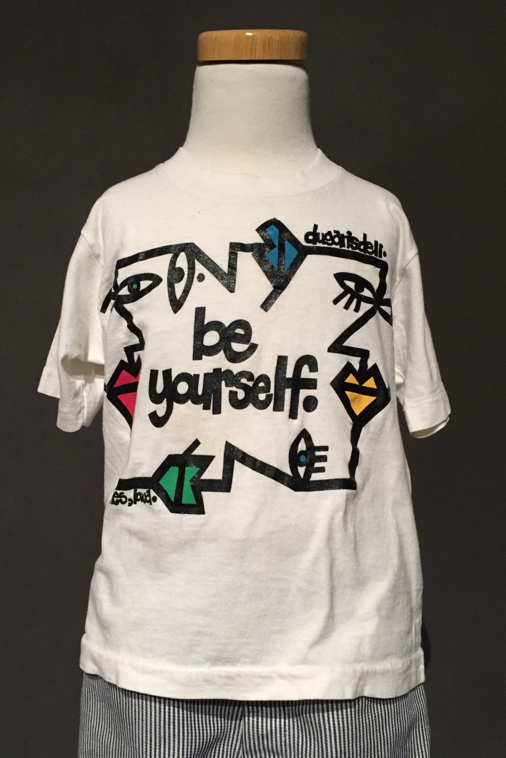 White short sleeve t-shirt with abstract geometric faces and black text reading: "Be Yourself" paired with black and white striped cuffed jeans