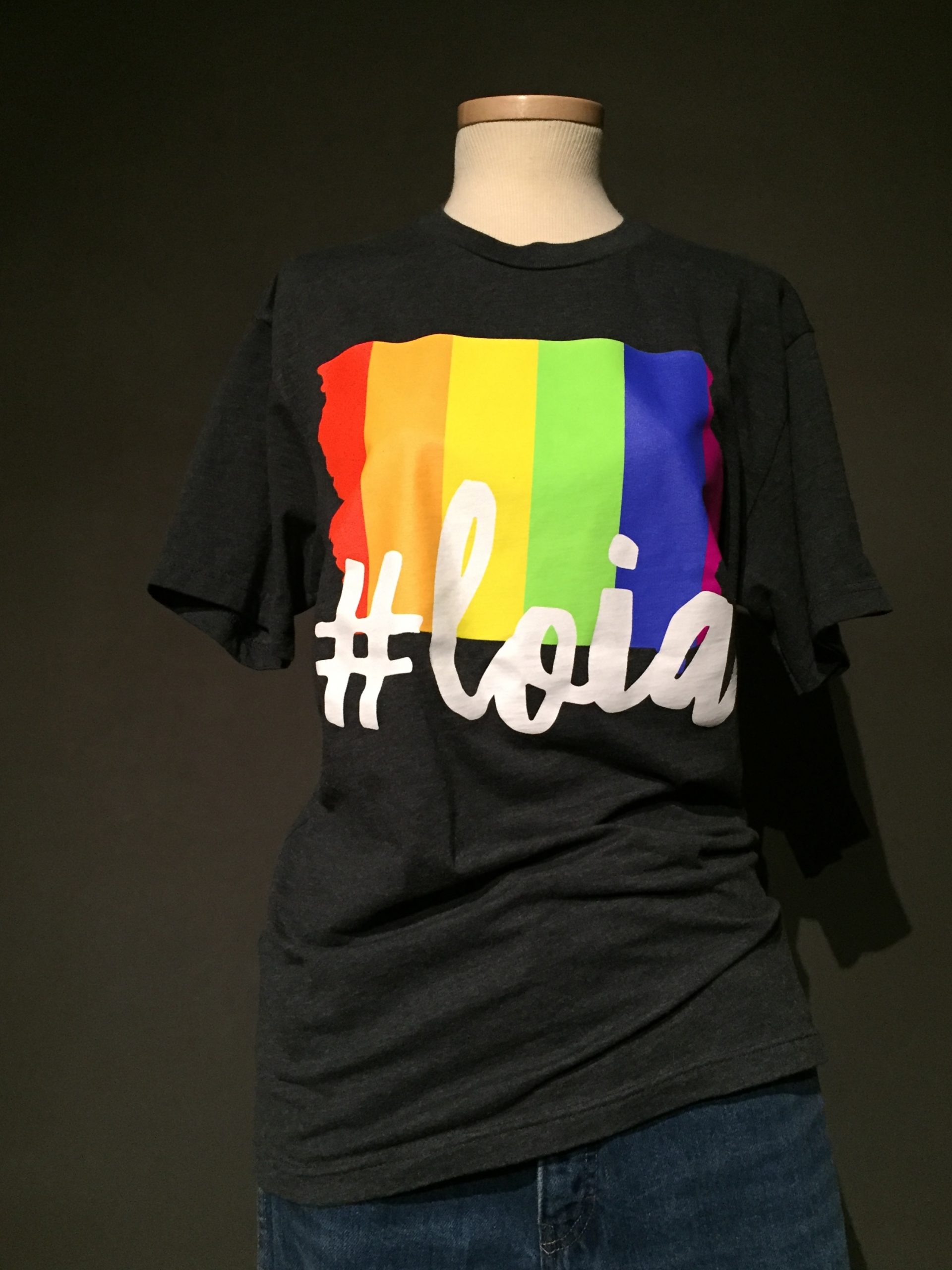 Black short sleeve t-shirt with outline of Iowa state filled in with rainbow, white lettering reading: "#LOIA" paired with dark straight leg jeans