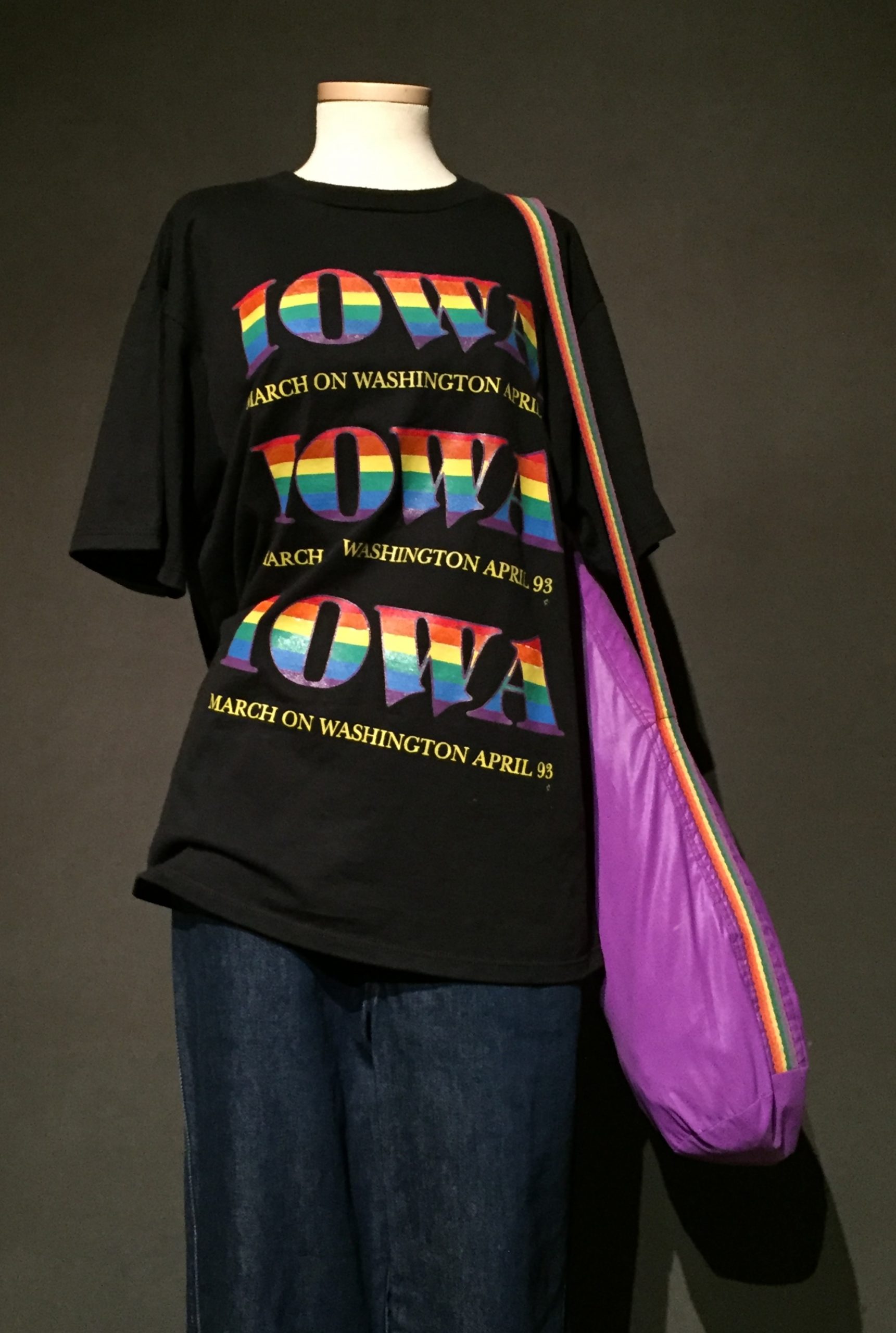 Black short sleeve t-shirt with rainbow lettering reading: "Iowa: March on Washington 93", cuffed denim pants, and purple bag with rainbow strap
