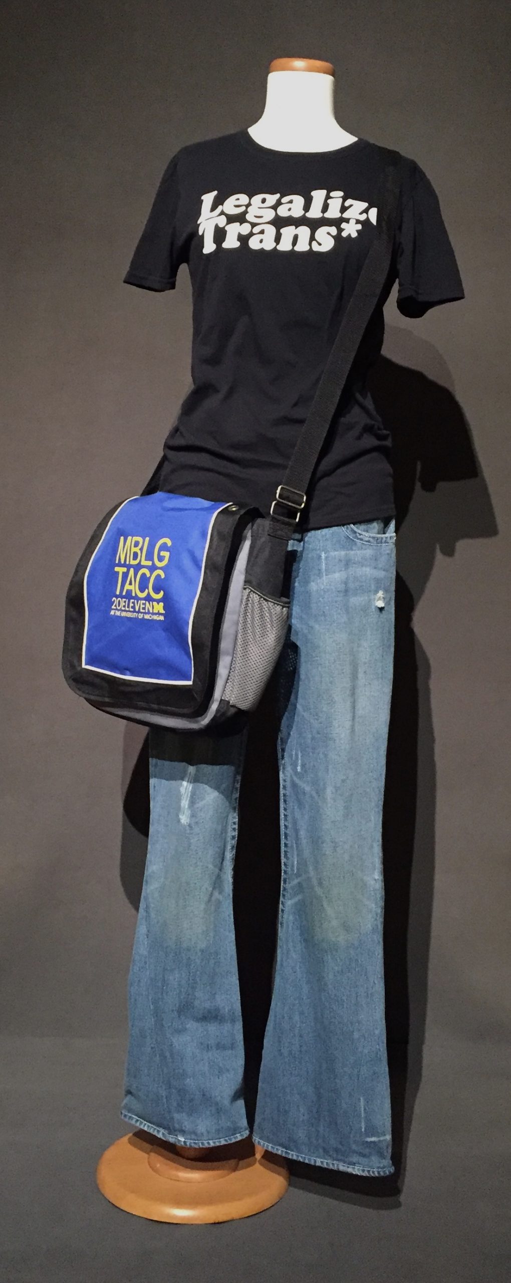 Black shirt with white text reading: "Legalize Trans*", flared jeans, and blue and black messenger bag with yellow text reading: "MBLG TACC 20ELEVEN at the University of Michigan"