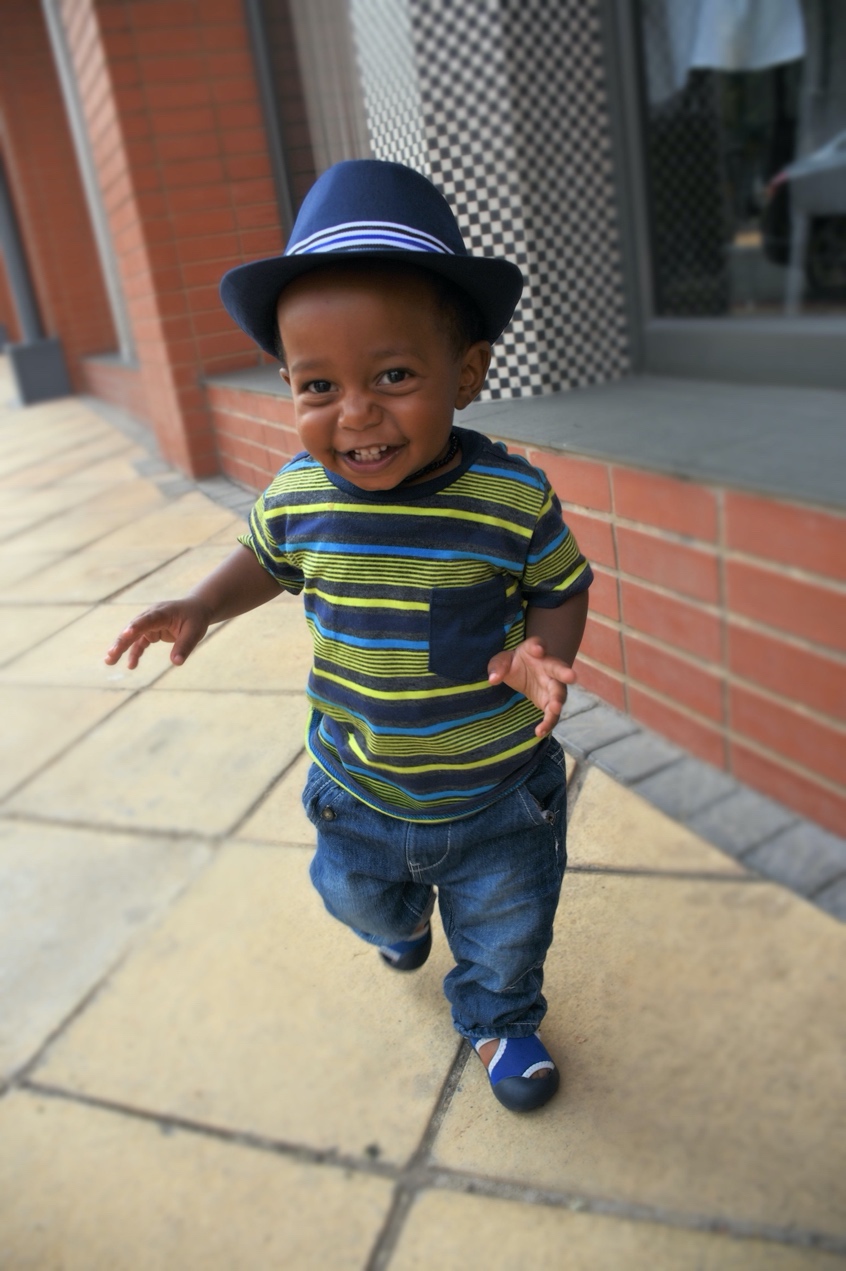 A smiling Black toddler in a hat walking toward the camera