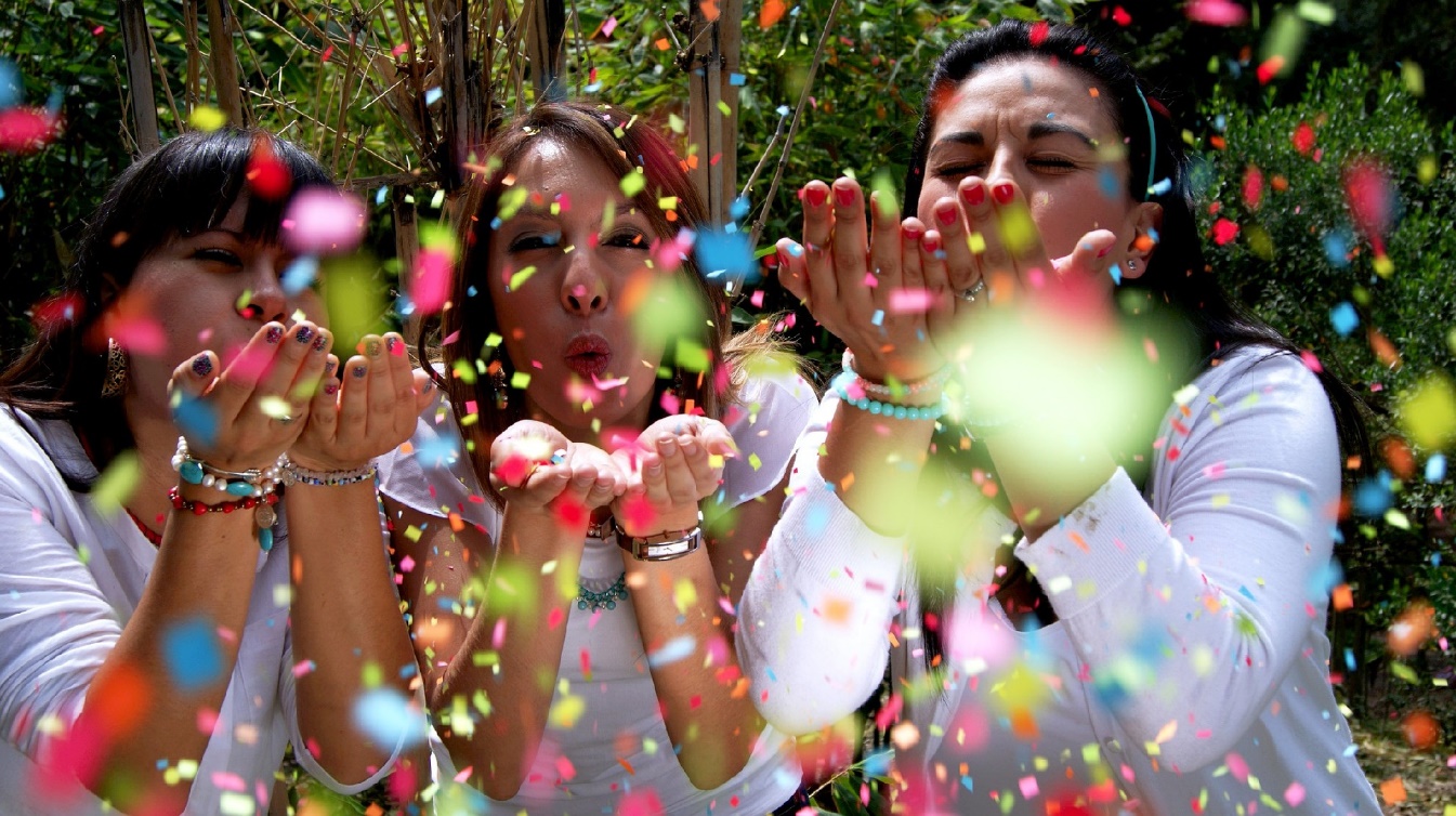 Photo of three women blowing confetti at the camera.