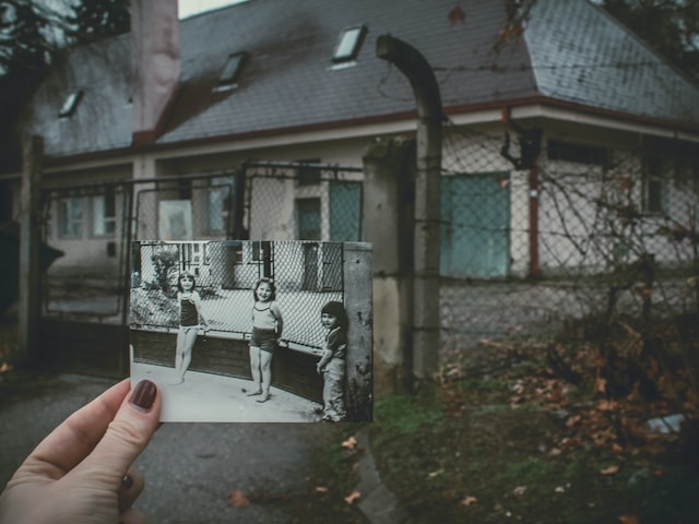 Photo of a person holding up a black and white photo of children against a dilapidated looking house.