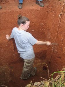 Amber standing is a soil pit, with red-orange colors, holding a soil knife.
