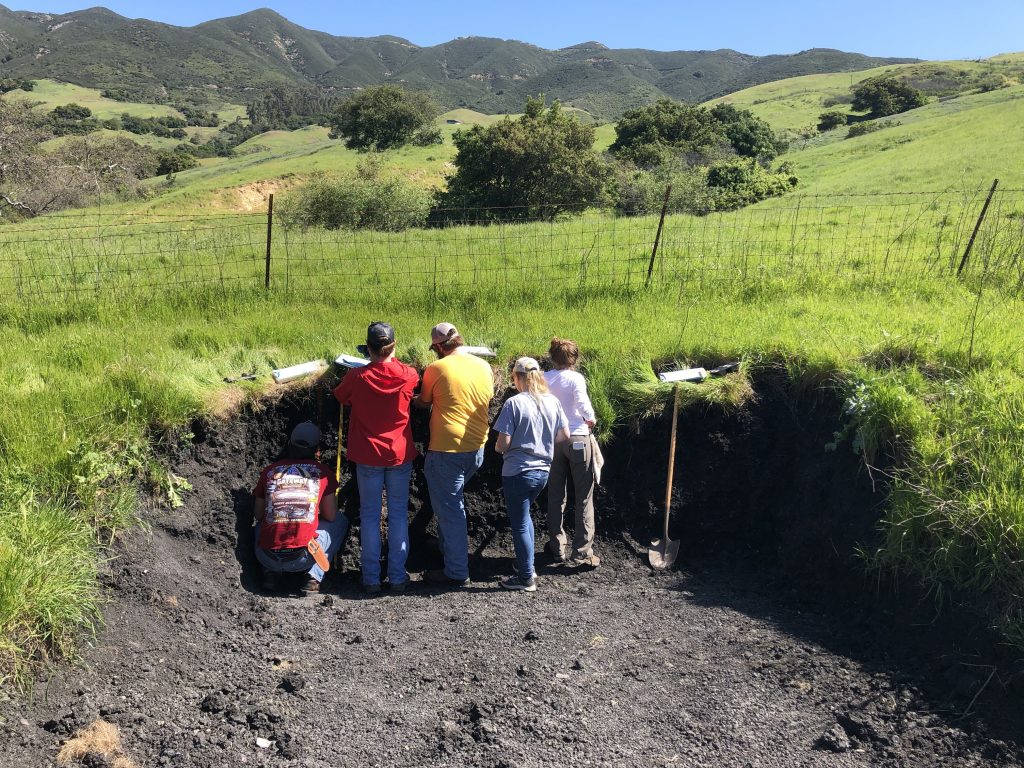 5 ISU soil judgers examining a soil face that is dug out of a rolling hill landscape several feet from a wire fence line. The background is hilly and has a blue clear sky.