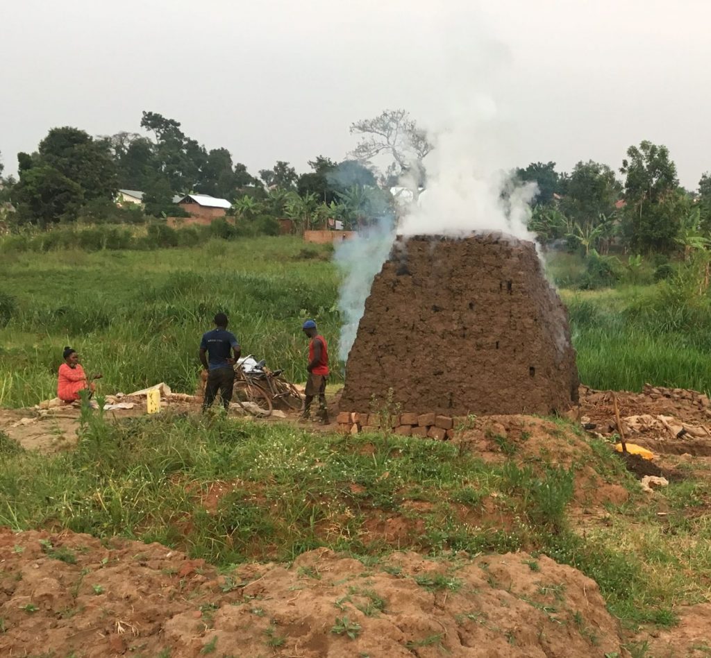 Clay is being harvested and formed into shapes to then be placed on in a tower around a fire to be made into bricks. The oven site has smoke coming out of the top.