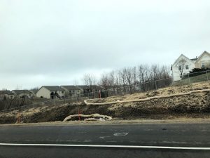 Exposed soil on the side of the interstate with straw waddles and straw place on bare soil. There are houses and tress with no leaves in the background. The sky is cloudy.
