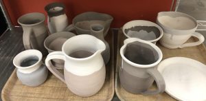 Assorted pitchers, vases, and a plate on two lunch trays with dull-colored clay materials that will be transformed in the kiln heating process into more durable goods with more vibrant colors