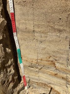 Soil pit with tape measure showing from 75 to 132 cm of depth, between 105 and 130, horizontal layers of different red and grey shades run across the pit. Finer textures appear more grey.