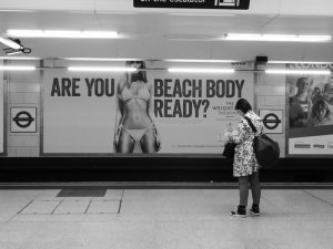 A woman standing in front of an ad in a subway station. The ad features a woman wearing bikinis with a catchline that says, "Are you Beach Body Ready"