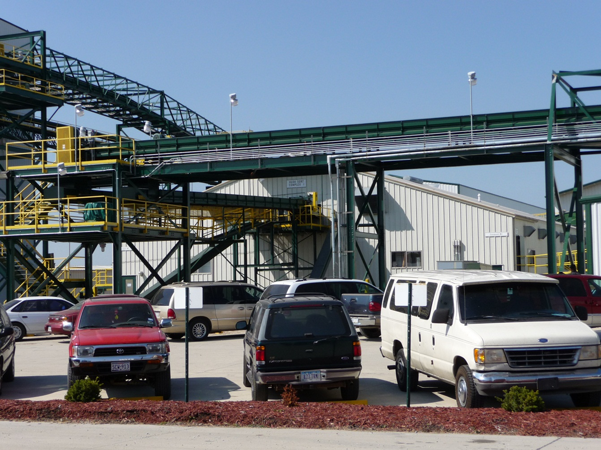 A parking lot with a factory beyond it. Conveyor belts can be seen moving corn.